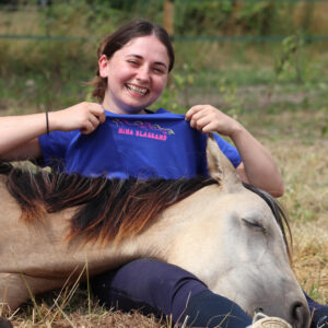 poulain isabelle couché sur sa cavalière qui porte un t-shirt d'équitation bleu en coton biologique avec une illustration engagée pour le bien-être équin