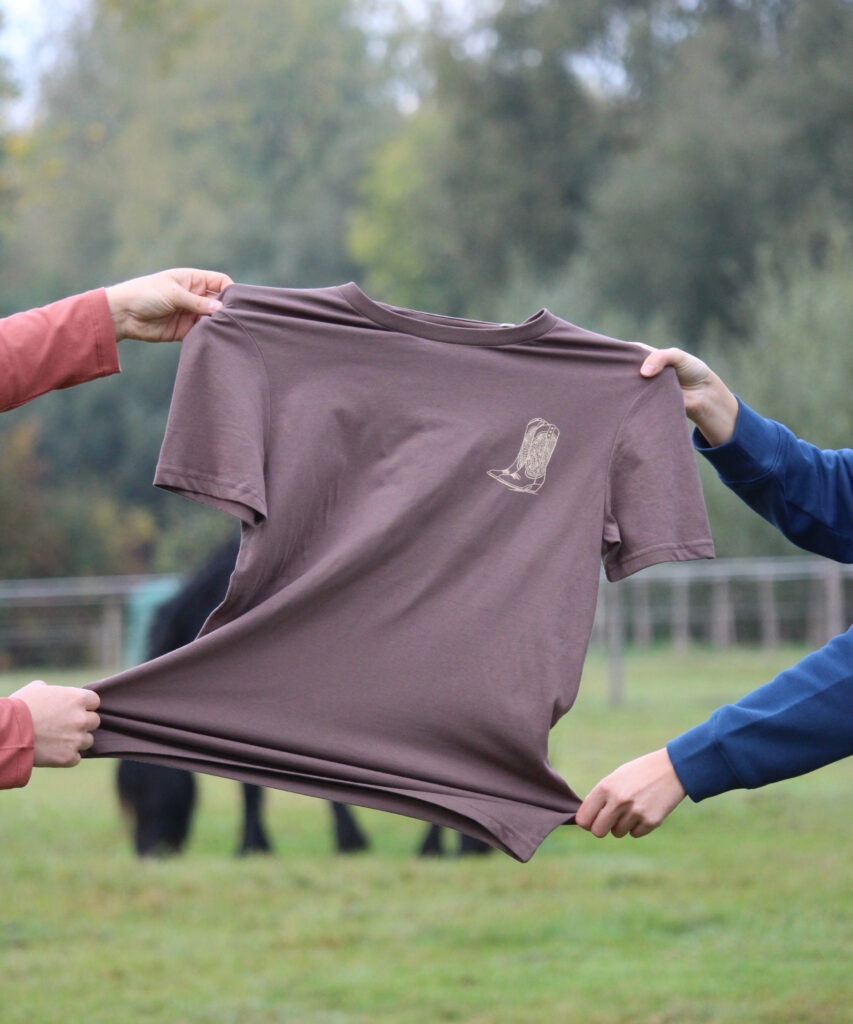 t-shirt d'équitation marron pour les cavalières western avec des bottes de cowgirl devant une prairie avec des chevaux