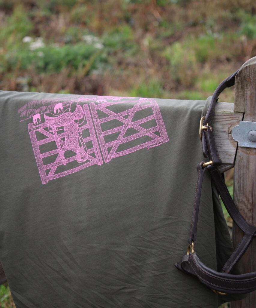 t-shirt d'équitation en coton biologique vert kaki avec une impression rose d'une selle western et d'un paysage du montana sur une barrière d'écurie avec un licol en cuir