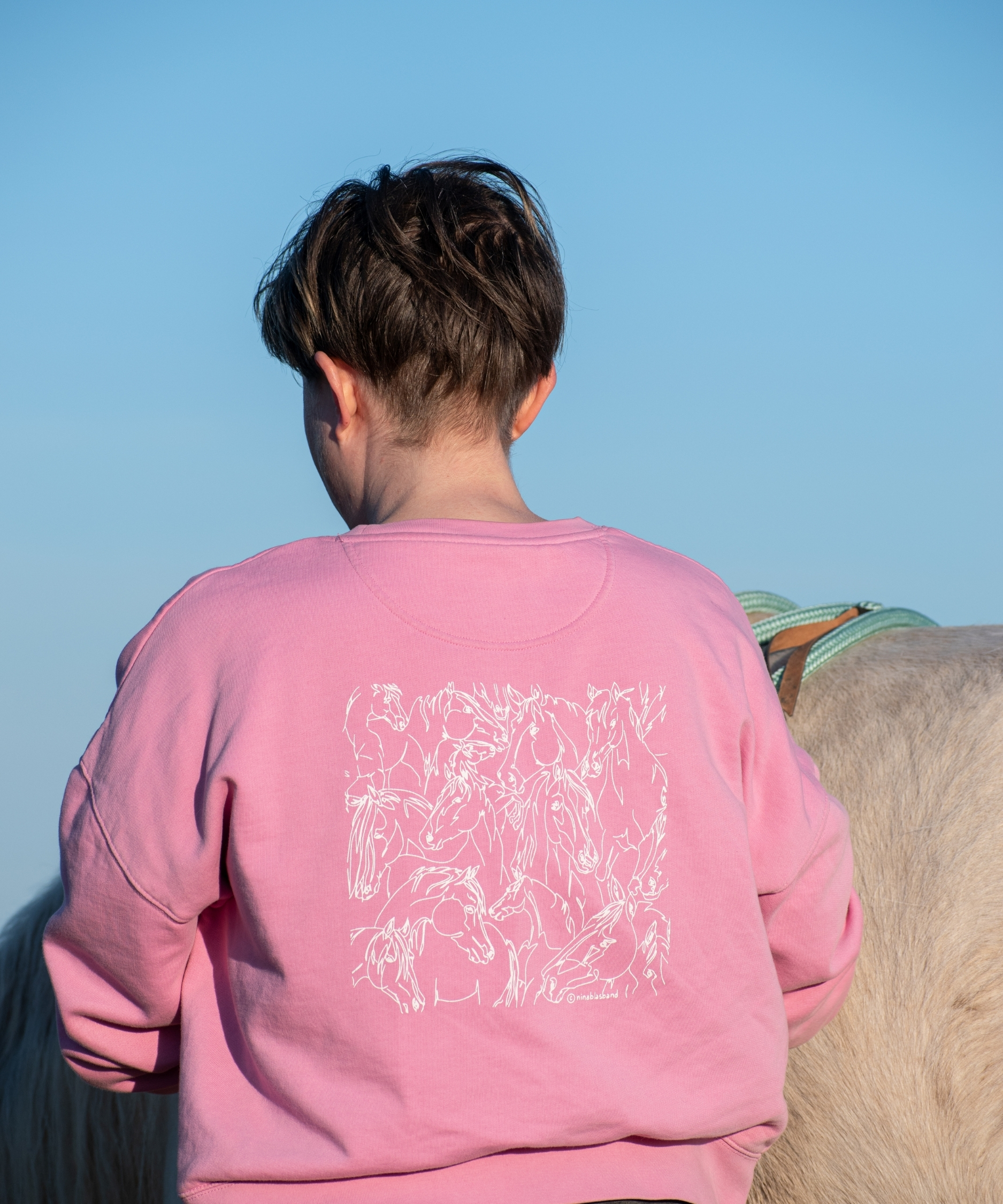 cavalière de dos avec un pull équestre rose en coton biologique illustré de chevaux