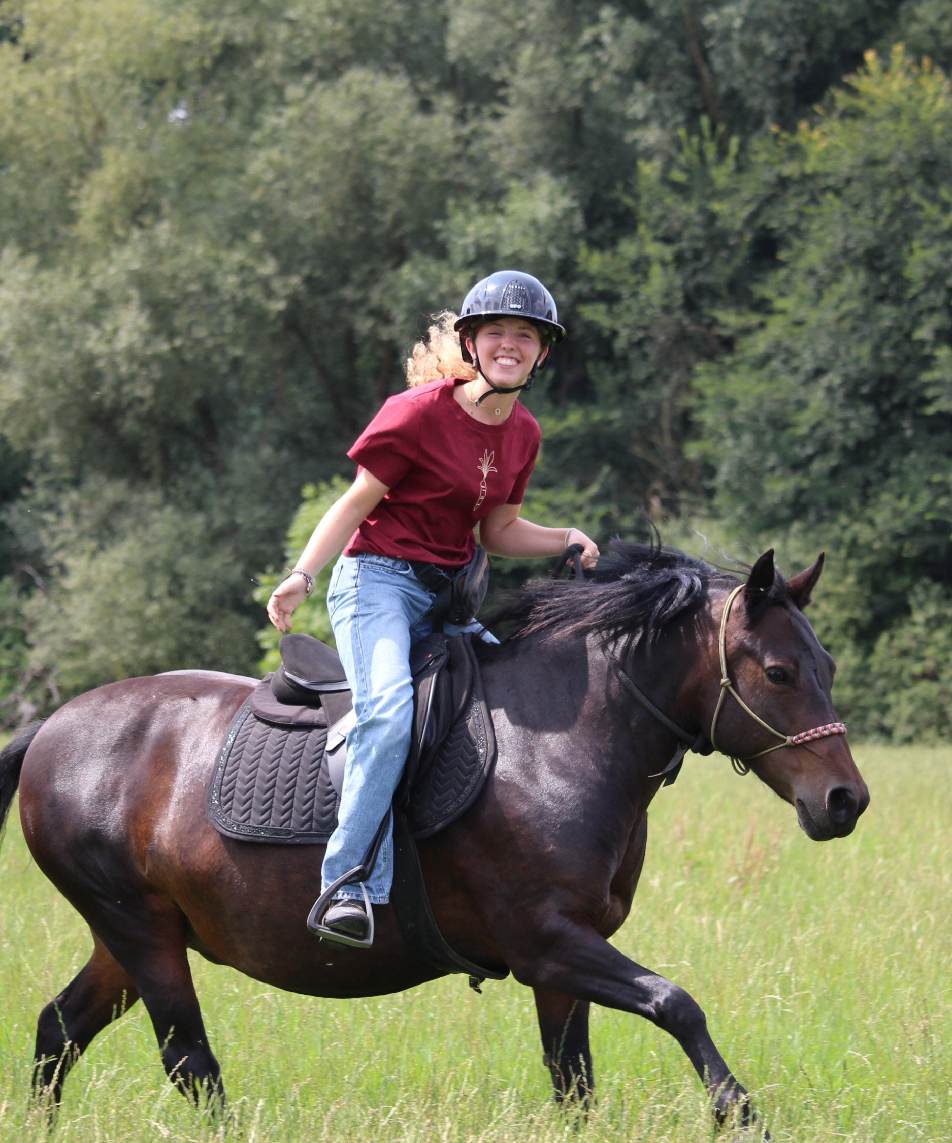 jeune cavalière au galop sur sa ponette avec un tapis de selle kentucky dans un champs en licol éthologique porte une bombe et un t-shirt équestre bordeaux en coton biologique avec une illustration de carotte