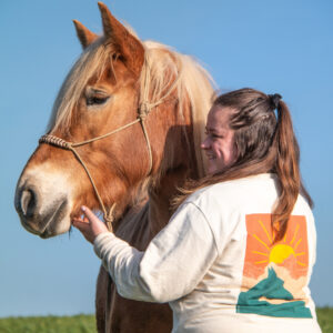 cheval haflinger qui pose avec sa cavalière portant un pull d'équitation en coton biologique avec une illustration de coucher de soleil